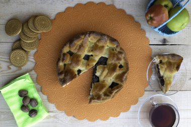 Turta, bir parça vişneli ve cevizli elmalı turta, bir fincan çay, sepetteki elmalar, yeşil peçetedeki cevizler ve masada kurabiyeler.