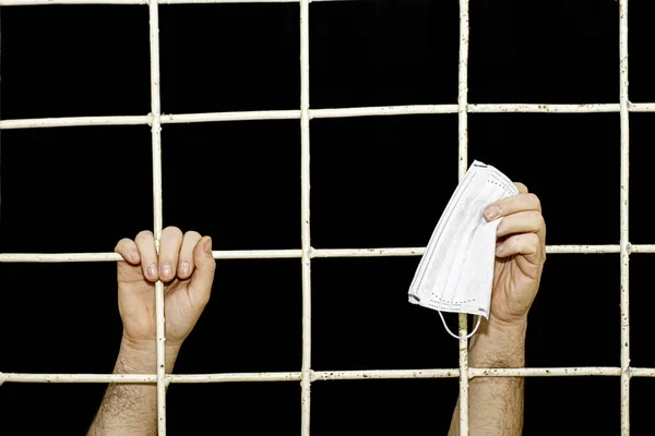 Concept of restriction during the pandemic and quarantine of a person s hand behind an iron bars in his hand medical hygiene mask — Stock Photo, Image