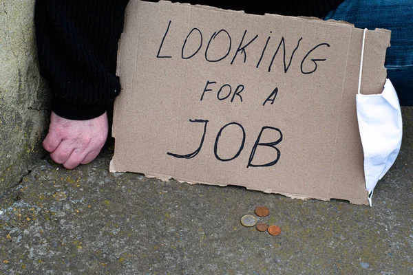 Concepto de desempleo y coronavirus desempleado sentado en el suelo junto a él una placa de cartón con la inscripción en busca de un trabajo, la máscara de higiene médica y monedas —  Fotos de Stock