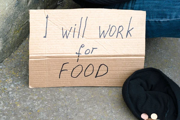 Unemployed man sitting on the ground holding the cardboard sign saying i will work for food next to is a hat with alms coins — Stock Photo, Image