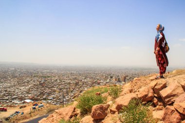 Kafası örtülü ve uzun ceketli bir kız turist İran 'ın Tabriz kentinin manzarasına karşı ayakta duruyor.