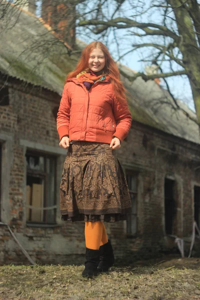 A happy red-haired young girl in a bright red jacket and pantyhose, in a skirt with floral patterns, poses against the backdrop of nature and the old village house.