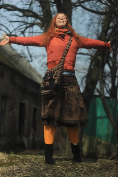 一个红头发 穿着鲜红色夹克和裤袜 穿着花纹裙子的快乐的小女孩 在大自然和村庄老房子的背景下摆出一副很高兴的架势 — 图库照片