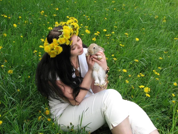 一个身穿白衣 一头长长的黑头发 头戴蒲公英花环的小女孩坐在一片绿油油的田野上 手里拿着一只可爱的小兔子 — 图库照片