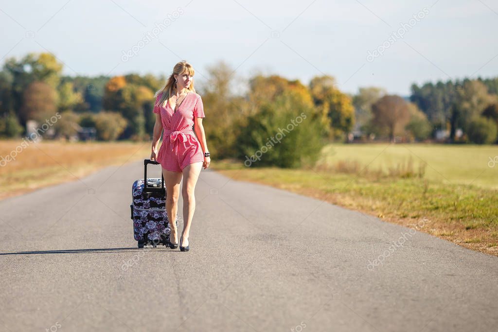young woman hitch-hiking on a road with luggage bag at the fields