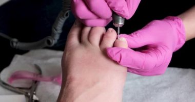 Pembe eldivenli pedikür uzmanı, pedikür salonundaki tırnak ve şalgam tırnaklarını matkapla kesiyor. Kozmetoloji kliniğinde profesyonel pedikür. Kuaförde ayaklar için hijyen.