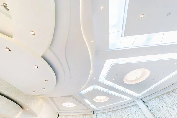 looking up on suspended ceiling with halogen spots lamps and drywall construction in empty room in apartment or house. Stretch ceiling white and complex shape.