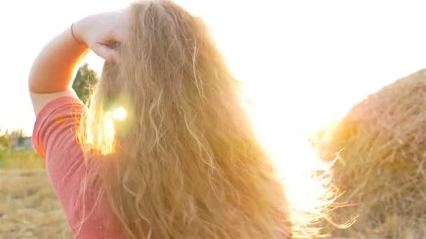 Ένα όμορφο κορίτσι στο ηλιοβασίλεμα προσαρμόζει τα μαλλιά της. Αργή κίνηση. Μια στοίβα του σανού — Αρχείο Βίντεο