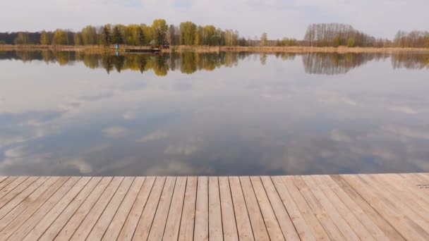 Pintoresco lago con un muelle de madera. Una cabaña de caza al otro lado. El cielo azul se refleja en la superficie del agua. La cámara avanza. Movimiento lento — Vídeo de stock