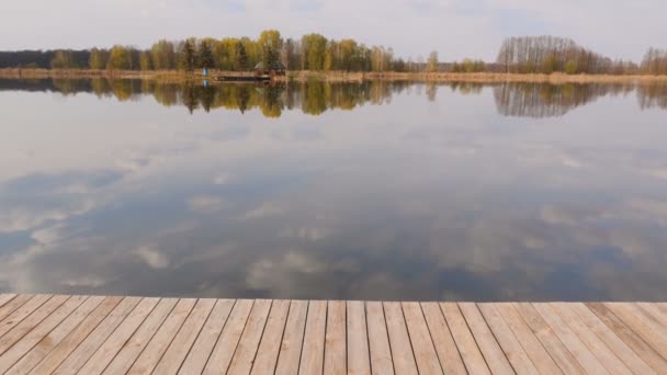 Träpir på sjön. Ensamt hus på andra sidan. Vackert soligt väder. Ljusblå himmel. Moln reflekteras i vattnet. Kamera i rörelse — Stockvideo