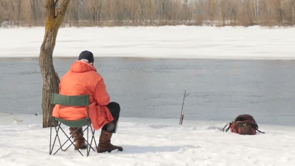 Человек на берегу ловит рыбу зимой. Живописное место вдали от людей. Поток воды посреди замерзшей реки — стоковое видео