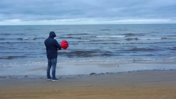 Человек в синей куртке открывает большой красный зонтик на берегу моря — стоковое видео