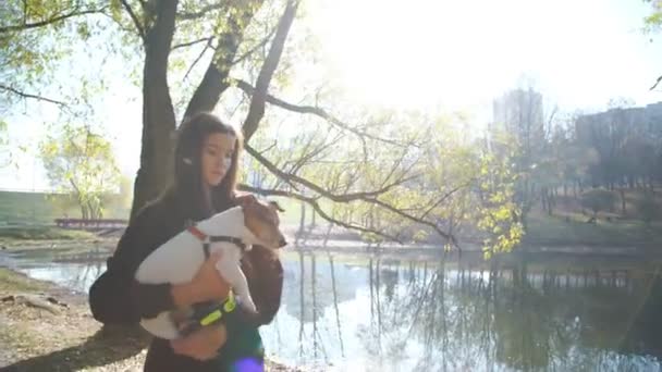 Chica camina en orilla del lago sosteniendo mascota en brazos contra la luz del sol — Vídeos de Stock