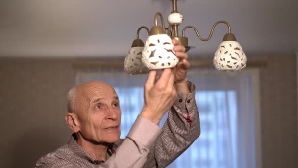 Концентрированный старик в клетчатой рубашке откручивает блестящую лампочку — стоковое видео