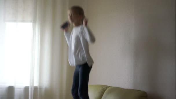 Девушка с толстой косой в белой рубашке и джинсах прыгает на диване — стоковое видео