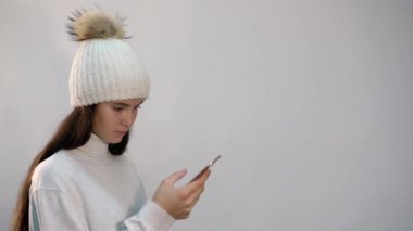 Beyaz şapkalı genç kız açık arkaplanda telefonla mesajlaşıyor.