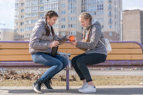 Saçları örgülü genç ve güzel kızlar termostan sıcak çay içerler. Şehir sokaklarındaki ahşap bankta otururlar.