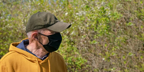 Homem velho em preto máscara facial descartável olha para árvores de jardim — Fotografia de Stock