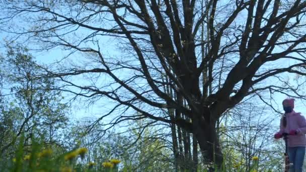 Школьница в маске проезжает на скутере мимо большого голого дерева — стоковое видео