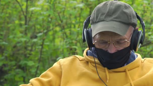 Hombre mayor en fideos máscara hablando sobre auriculares en el jardín — Vídeo de stock