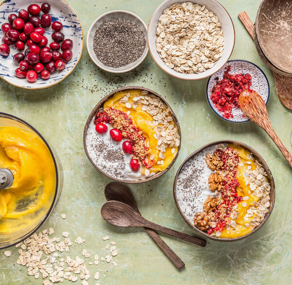 Healthy clean breakfast with smoothie bowl with mango and  tropical fruits , chia seeds yogurt pudding and cranberries, nuts, oatmeal topping in coconut shells with spoon, top view.