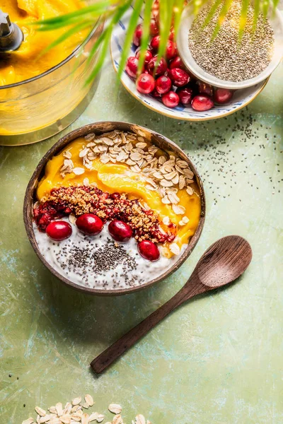 Smoothie bowl with mango and  tropical fruits , chia seeds yogurt pudding and cranberries, nuts, oatmeal topping in coconut shells with spoon, top view. Healthy clean breakfast food. Summer vegan diet