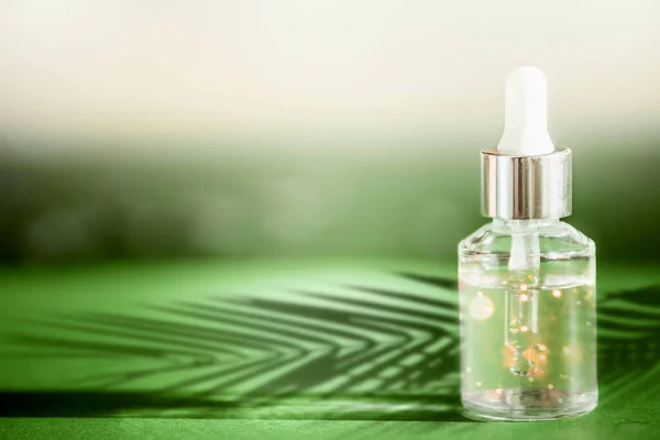 Transparent cosmetic bottle with pipette standing on green table with palm leaves shadow. Facial skin care concept. Natural vegan cosmetic. Serum or skin essential oil. Modern beauty trend.