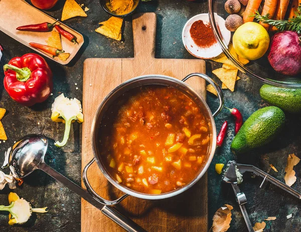 Mexican vegetarian  bean soup in cooking pot with ladle on rustic kitchen table ingredients and cutting board, top view. Vegan or vegetarian healthy food concept