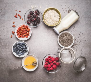 Süper yiyecekli çeşitli sağlıklı kahvaltı malzemeleri: chia tohumları, goji üzümleri, yulaf ezmesi, taze böğürtlen, bal ve süt veya gri arka planda yoğurt, üst görünüm, çerçeve