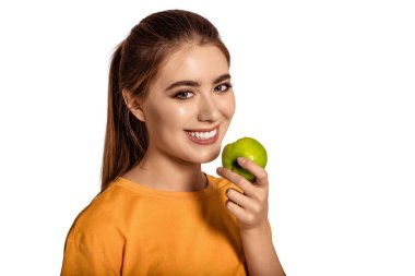 Güzel, genç bir kız elinde yeşil bir elma tutuyor ve dişleriyle gülümsüyor. Kar beyazı, mutlu bir gülümseme. Ağız bakımı. Diyet, vejetaryenlik. Sağlıklı yaşam tarzı.