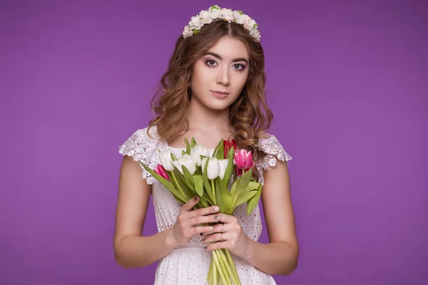 A beautiful young woman stands on a purple background, wearing a white dress and a wreath of flowers on her head, and a bouquet of tulips. International Women\'s Day, March 8.