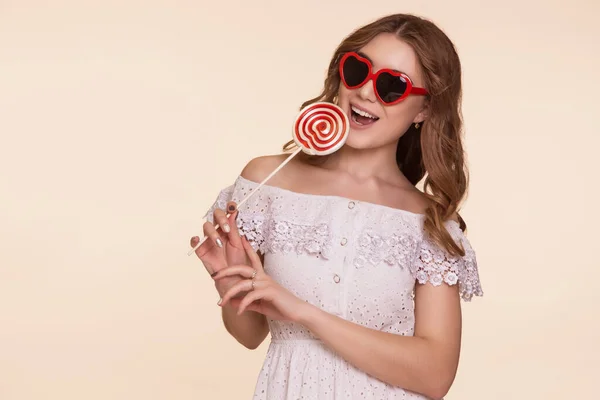 腕と眼鏡の中に赤と白のキャンディで立っている美しい若い女性の心は ベージュの背景 白いドレスの少女ロリータ コピースペース — ストック写真
