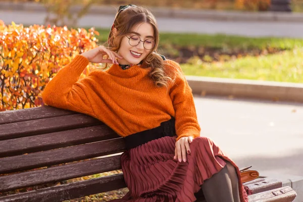 美しい若い女性が10月の秋にベンチに座っています 女の子は眼鏡をかけて丸い服を着ている 生徒の先生が休んで公園を歩いてる — ストック写真