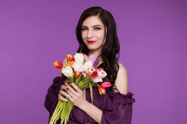 Güzel, genç bir kadın, esmer, mor bir arka planda duruyor, hava elbisesi ve bir buket lale ile. Uluslararası Kadınlar Günü, 8 Mart.