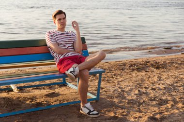 Yakışıklı, genç bir adam, beyaz çizgili gömlekli ve şortlu bir adam nehir kenarındaki bir bankta oturuyor. Tatil köyündeki sahil. Deniz havasını içine çek, iyot eksikliği..