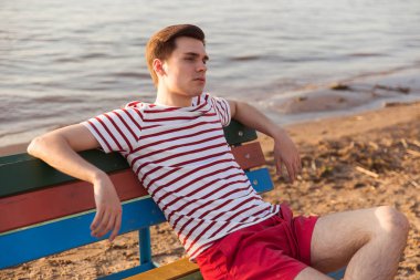 Yakışıklı, genç bir adam, beyaz çizgili gömlekli ve şortlu bir adam nehir kenarındaki bir bankta oturuyor. Tatil köyündeki sahil. Deniz havasını içine çek, iyot eksikliği..