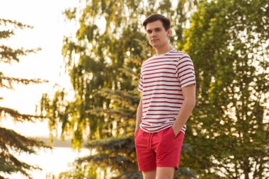 Yakışıklı, genç bir adam, beyaz çizgili gömlekli ve şortlu bir adam ağaçların arkasında duruyor. Parkta. Temiz havada yürü. Yola devam etmek için. Model pozu. Milyonlarca, Z jenerasyonu.