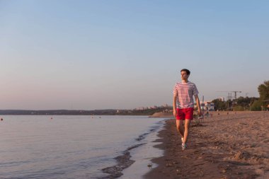 Yakışıklı, genç bir adam, beyaz tişörtlü, çizgili ve şortlu bir adam nehir kenarında yürüyor, deniz kenarında. Otelde yürüyordum. Sahilde dinlen..