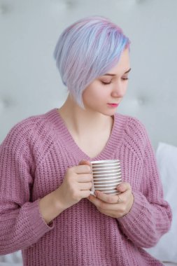 Mavi saçlı güzel bir kadın, yatak odasında, mor bir kazak giymiş, elinde sıcak kahve ya da çay bardağı tutan bir yatakta oturuyor..