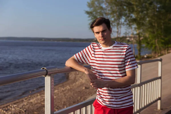 A handsome, young guy, a man in a white striped shirt is leaning on a bridge and is looking into the distance. Life style, copy space. Character from old movies.