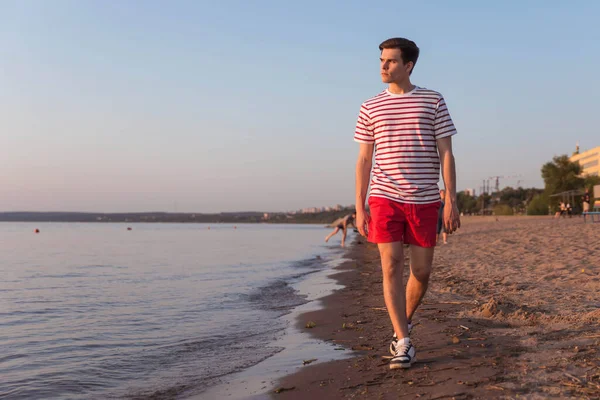 A handsome, young guy, a man in a white T-shirt with stripes and shorts is walking along the embankment of the river, the sea. Walking at the resort. Rest by the beach. The character of the old movie.