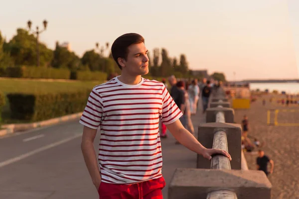 A handsome, young guy, a man in a white striped shirt is leaning on a bridge and is looking into the distance. Embankment, beach resort. Life style. Character from old movies.
