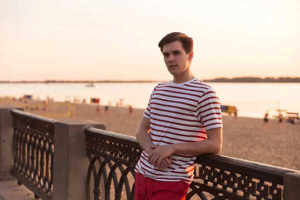 A handsome, young guy, a man in a white striped shirt is leaning on a bridge and is looking into the distance. Embankment, beach resort. Life style. Character from old movies.