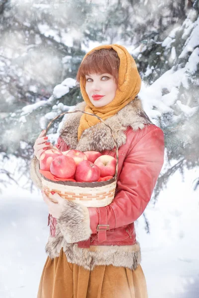 美しいスラブ人の女性は 冬には 針葉樹の木の背景にあるスカーフで 赤いリンゴの入ったウィッカーバスケットを保持しています 寒いロシアの冬 — ストック写真