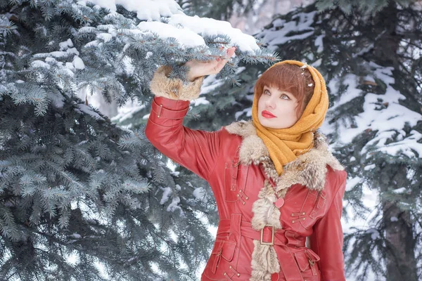 美しいスラブ人の女性は 冬には 頭の上のスカーフで 針葉樹の木の背景を背景に 遠くを見ています ロシアの冬 大きな赤い唇 雪が降る — ストック写真