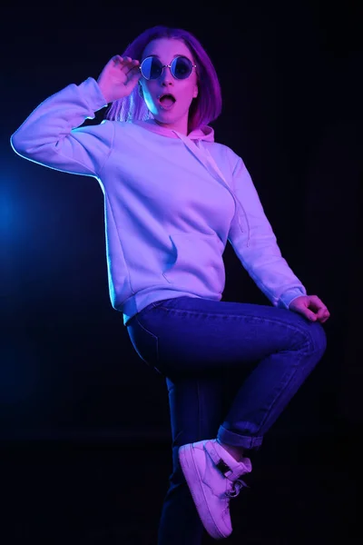 A woman with colored hair, an informal female drowns on a black background in a club, in neon lights and sunglasses, dancing. The girl is dressed in a lilac sweatshirt, sweater. Night life.