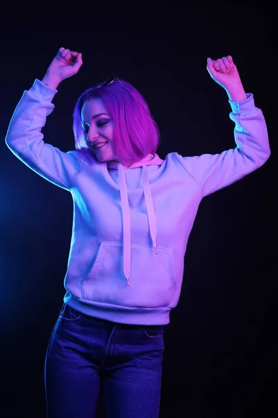 A woman with colored hair, an informal woman drowns on a black background in a club, in neon light. The girl is dressed in a lilac sweatshirt, sweater. Night life.