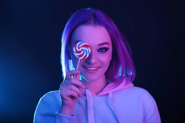 A woman with colored hair, an informal woman drowns on a black background in a club, in neon light. Holding a lollipop. Night life.