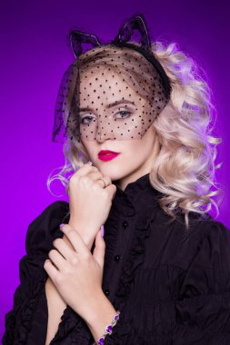 Güzel, genç, sarışın, siyah retro bluzlu, kedinin kulaklarında ve siyah dantelli peçeli bir kadın. Modaya uygun bir portre. Mor arka planda kadın kedi