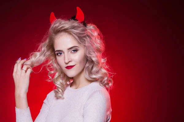 To twist a curl of hair on a finger. Portrait of a beautiful blonde woman with a cunning look and horns on her head. The dress for the Halloween party. Evil demon girl is the devil.